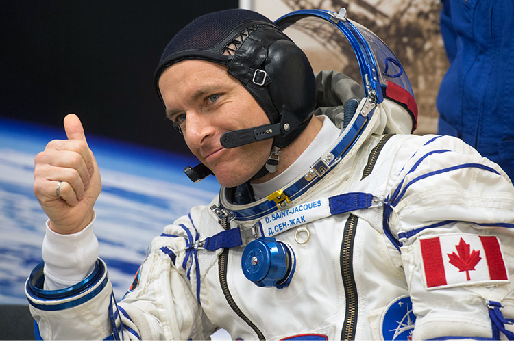 L’astronaute canadien David Saint-Jacques regarde à notre gauche en souriant et en faisant le signe du pouce levé. Il porte une combinaison spatiale de cosmonaute russe Sokol KV2, sans les gants et avec la visière de casque relevée. 
